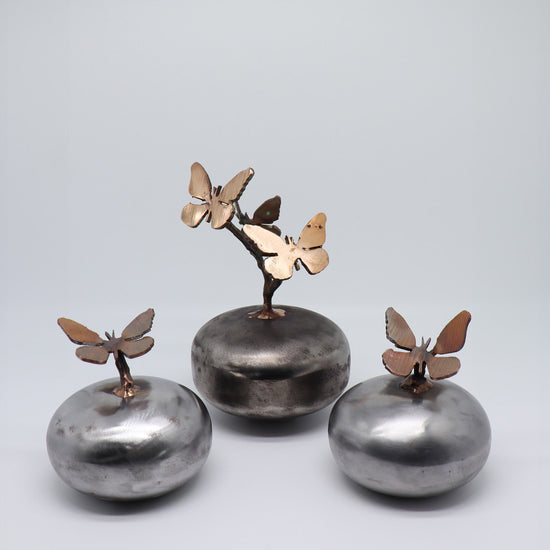 Familie set van 3 mini urnen van JEART met bronzen vlinders, op ronde stalen voetstukken. Symboliseert transformatie en verbondenheid. De combinatie van delicate bronzen beelden en stevige stalen bollen biedt een stijlvolle herdenkingsplek voor dierbaren.